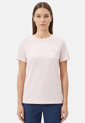 Giovane donna con capelli castani lisci, che indossa una maglietta a maniche corte rosa chiaro e pantaloni blu scuro, in piedi su uno sfondo semplice.