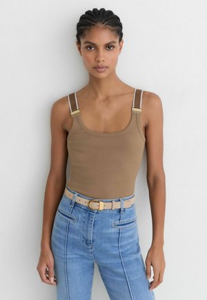 Frau mit geflochtenem Haar, die ein beige geripptes Tanktop mit Schnallen-Trägern, hellblaue hoch taillierte Jeans und einen beigen Gürtel mit goldener Schnalle trägt.