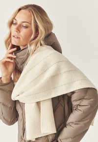 Krēmkrāsas adīta wraps ar svītrainu rakstu, p�ārklāts pār brūnu pūkainu jaku. Wrapam ir riboti akcenti un mīksta tekstūra.