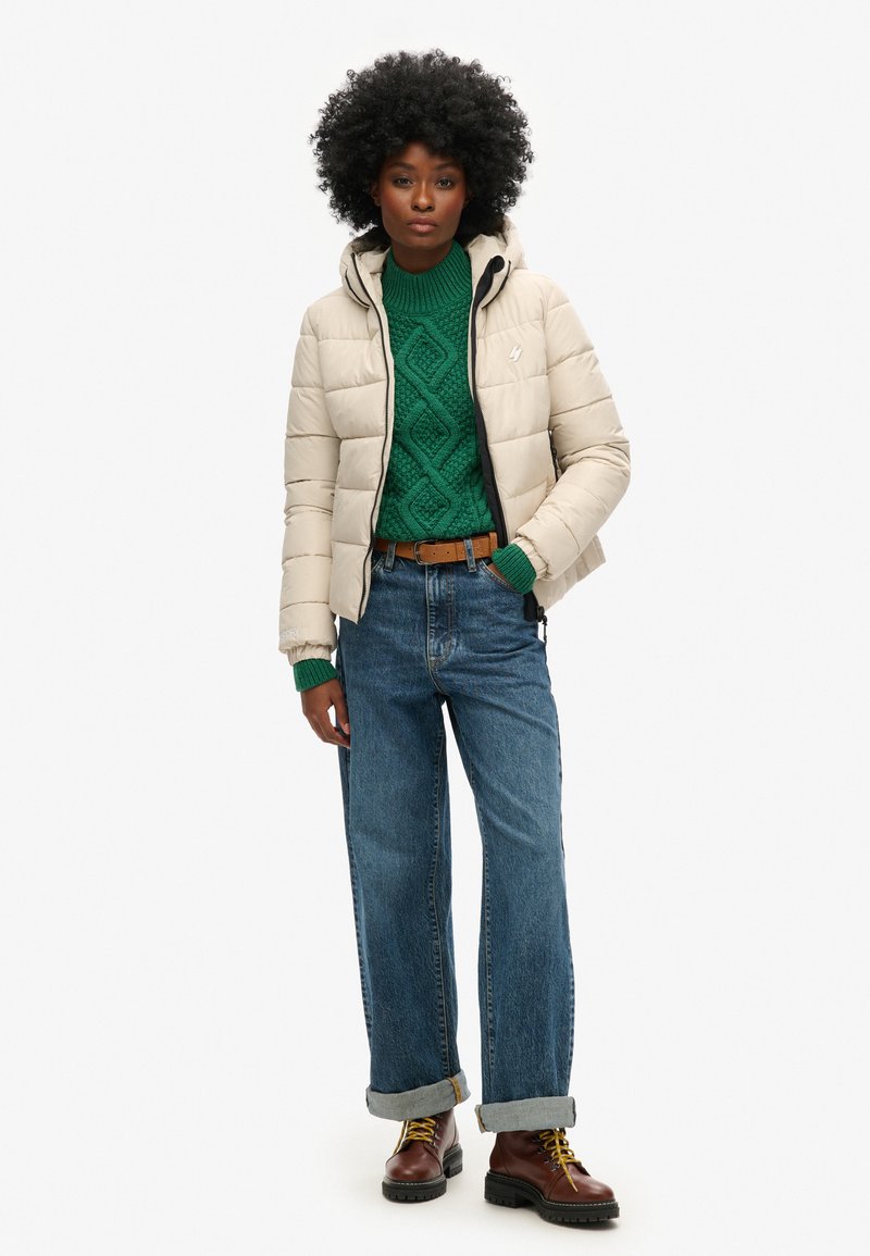 Beżowa kurtka puchowa z kapturem, w połączeniu z zielonym swetrem z warkoczem, spodniami dżinsowymi o wysokim stanie w kolorze niebieskim oraz brązowymi butami za sznurówki.