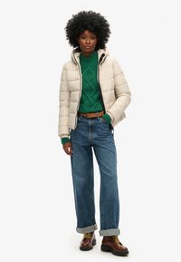 Beige vadderad jacka med huva, grön stickad tröja med flätmönster, blå vidbyxade jeans och bruna snörskor.