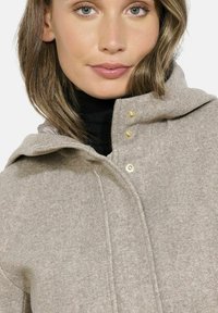 Cappotto in misto lana beige con cappuccio, dotato di bottoni a pressione dorati, una finitura testurizzata e una chiusura con zip. Sotto è presente un dolcevita nero.