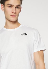 T-shirt bianco in cotone con colletto rotondo, maniche corte e logo nero sulla parte sinistra del petto. Texture liscia e design semplice.