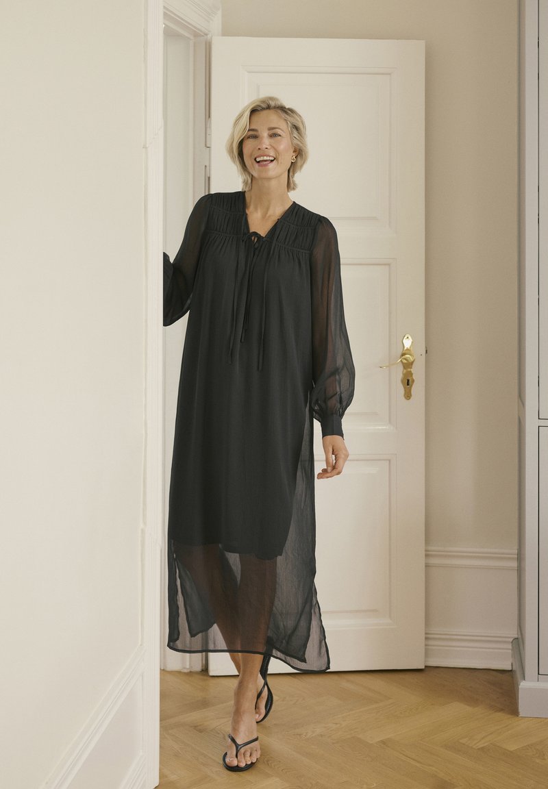 Schwarz, durchscheinendes Kleid mit langen Ärmeln, gerafften Details und einer Schleife am Ausschnitt. Fällt bis zur Mitte der Wade und hat eine entspannte Silhouette.