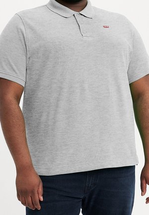 Mężczyzna noszący jasno szarą koszulkę polo z krótkim rękawem z małym czerwonym logo Levi's na piersi oraz ciemnoniebieskie dżinsy.