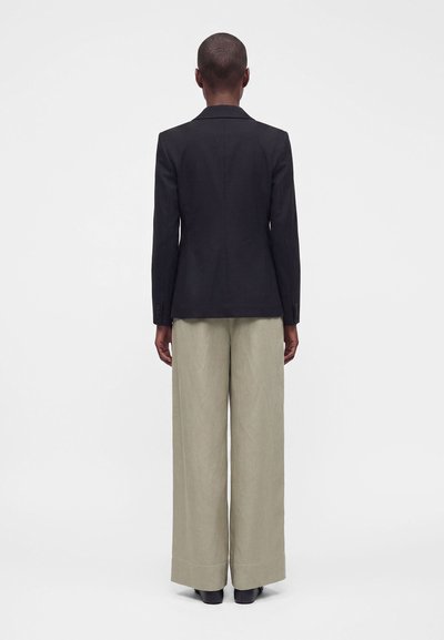 Personne aux cheveux courts portant un blazer noir et un pantalon beige large, debout, dos à un fond clair uni.