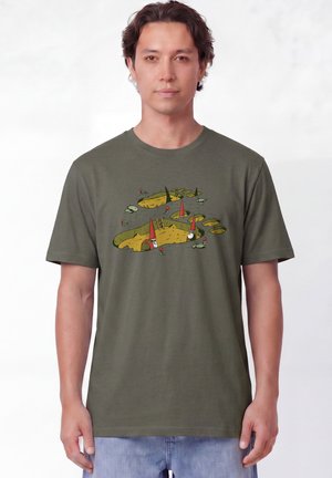 Homme portant un t-shirt vert olive avec des lutins en dessin animé creusant des tunnels et grimpant des échelles parmi des rochers et des champignons.