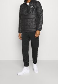 Zwarte gewatteerde pullover jas met een halve ritssluiting, voorzak, en een wit logo. Gecombineerd met zwarte joggingbroeken en witte sneakers.