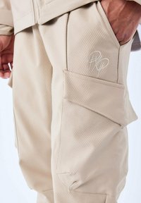 Beige cargobroek met verticale ribbeltextuur, voorzien van een geborduurd logo op de zak en een zijzak voor extra opbergruimte.