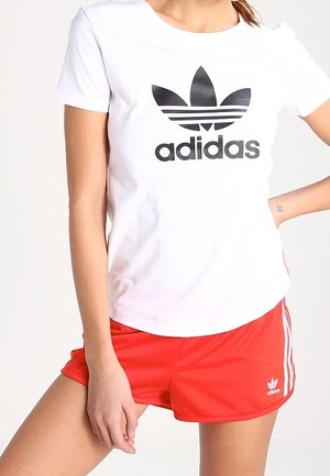 Frau trägt ein weißes T-Shirt mit Adidas-Logo und rote Adidas-Trainingsshorts mit weißen Streifen, posiert mit einer Hand in der Hüfte.