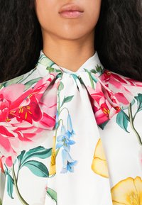 Blusa floreale in tessuto leggero con sfondo bianco e fiori rossi, blu e gialli, e dettaglio fiocco nel collo.