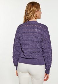 Maglione lavorato a maglia viola con un motivo in pizzo testurizzato, polsini e orlo a coste, visto da dietro, abbinato a pantaloni bianchi.
