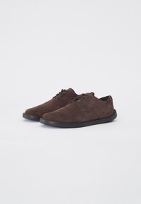Une paire de chaussures en suede marron foncé à lacets avec des semelles noires sur un fond blanc uni, présentée à un léger angle.