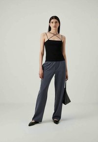Woman stands wearing black strappy sleeveless top, loose gray pants, black pointed flats, and holding small black handbag.