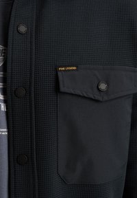 Temno modra teksturirana jopica z vafeljastim vzorcem, ki ima črne gumbe, žep na prsih in oznako PME Legend v rumeni barvi.