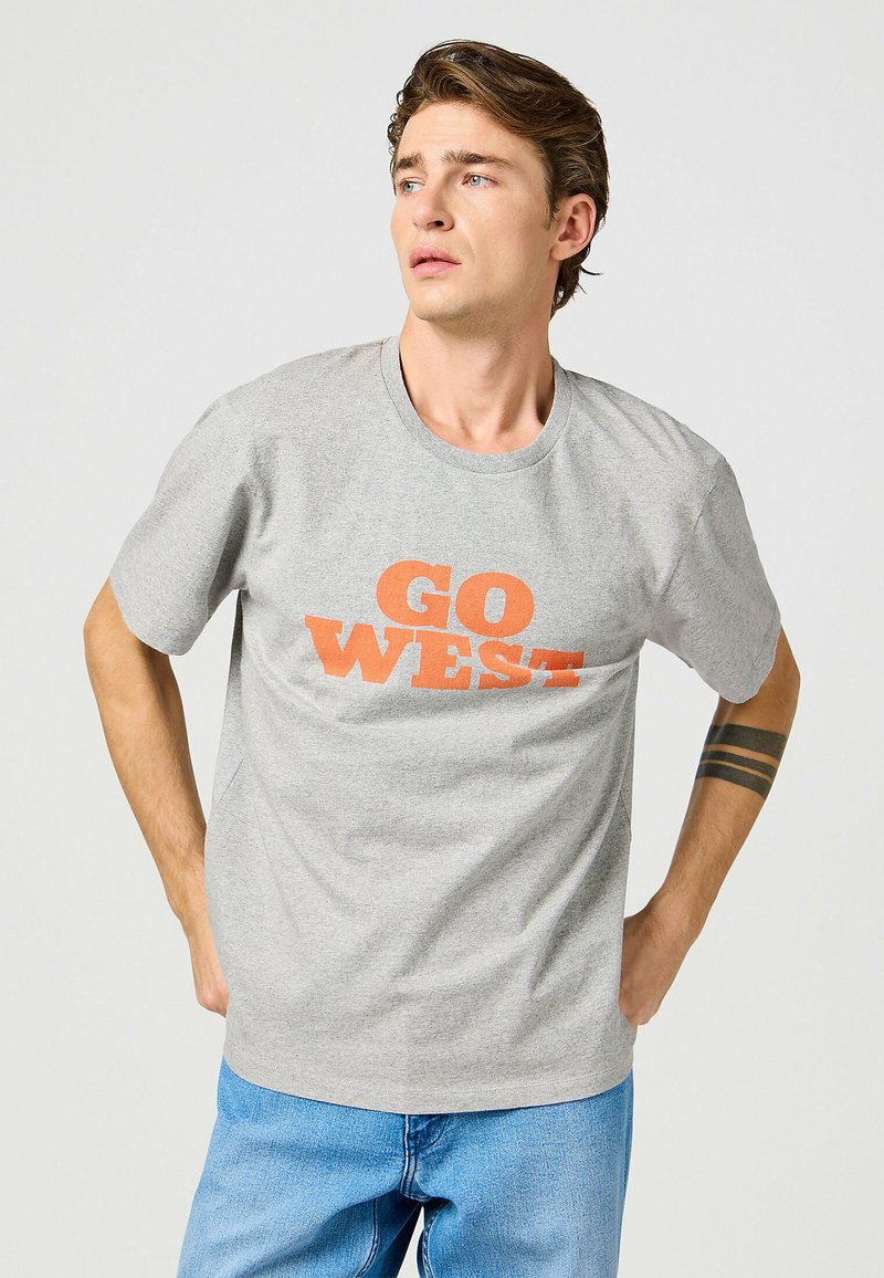 Camiseta de algodón gris con texto en bloque naranja que dice "GO WEST". Mangas cortas y cuello redondo. Exhibida en un modelo.