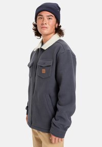 Tummansininen fleece-takki, jossa valkoinen tekoturkistakki, vetoketjukiinnitys, kaksi rintataskua ja vaalea logopatch; päällä tummansininen pipo.