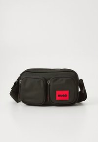 Kompaktna črna pasna torbica z dvema sprednjima žepoma na zadrgo, nastavljivim trakom in rdečo nalepko z napisom "HUGO" na desnem žepu.