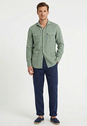 Hombre con camisa verde claro abotonada, pantalones azul marino y espadrilles azul marino, de pie sobre un fondo blanco liso.
