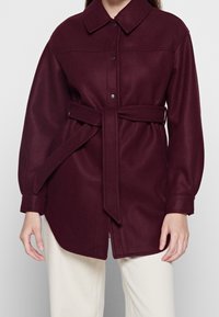Frau in einer burgunderfarbenen Wolljacke mit langen Ärmeln und Gürtel über weißen Hosen, die vor einem neutralen Hintergrund steht.
