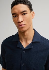 Marineblauw kortemouwen shirt van een gestructureerde stof, voorzien van een kraag en een drieknopenlijst. Subtiele horizontale lijnen in het materiaal.