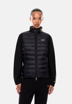 Vrouw die een zwarte EA7 Emporio Armani gewatteerde bodywarmer draagt over een zwarte top met lange mouwen en een zwarte broek tegen een witte achtergrond.