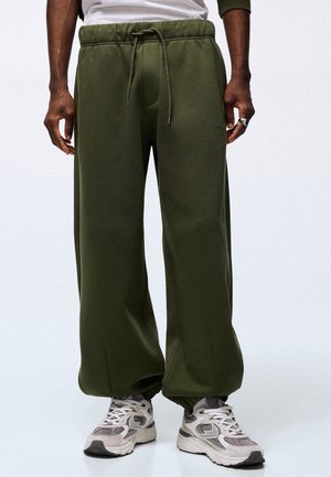 Homme portant un pantalon de survêtement large vert olive avec taille à cordon et des baskets blanches et grises sur un fond clair uni.