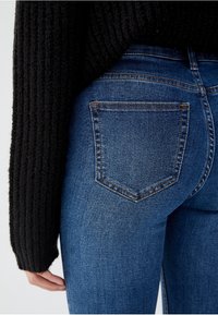 Jeans de tiro alto en azul con un diseño ajustado, que presentan un bolsillo trasero y un ligero desgaste. La modelo lleva un suéter negro de canalé.