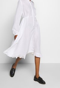 Biała lniana sukienka z guzikiem z przodu, długimi rękawami i falbaną u dołu, wcięta w talii z dopasowanym paskiem. Nosić ze czarnymi loafersami.