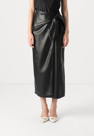 Vrouw draagt een zwarte rok van kunstleer met overslag, gestrikt in de taille, gecombineerd met witte pumps met spitse neus en metallic details.