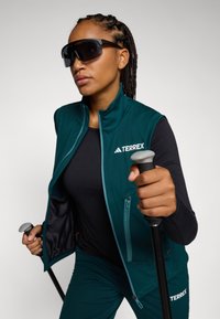 Türkise TERREX Weste über einem schwarzen Langarmshirt, hält Trekkingstöcke. Mit großen dunklen Sonnenbrillen und einem Reißverschlussdetail.