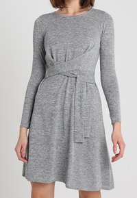 Grå långärmad klänning gjord av mjukt, strukturerat tyg. Har en figurnära bodice med en vriden midja och en utställd kjol.