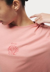 Rosa bomullströja med ett skalldesign och texten "La mer". Tyget är mjukt med en slät textur; ärmarna är lätt rullade.