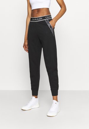 Mujer con joggers negros de Calvin Klein con cinturilla con marca y rayas, top corto blanco y zapatillas blancas, de pie con una mano en el bolsillo.