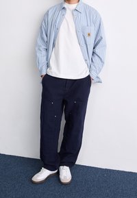Světlomodrá proužkovaná košile na knoflíky přes bílé tričko, v kombinaci s tmavě modrými volnými kalhotami a bílými teniskami s béžovými detaily.