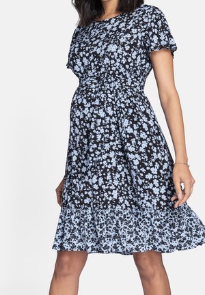 Kobieta ubrana w czarną i jasnoniebieską kwiatową sukienkę ciążową z krótkimi rękawami i falbaną na dole, stojąca na tle jednolitego tła.