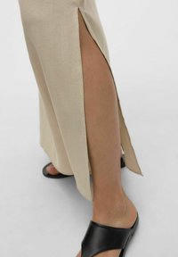 Beige dlouhá šaty s bočním rozparkem, vyrobené z hladkého materiálu. Nosí se s černými sandály, které mají minimalistický design a otevřenou špičku.