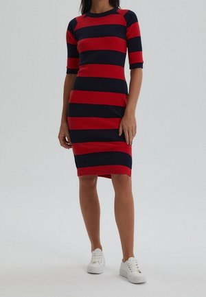 Donna che indossa un vestito a righe orizzontali rosso e blu navy, di lunghezza al ginocchio, con maniche a tre quarti e sneakers bianche, in piedi su uno sfondo neutro.
