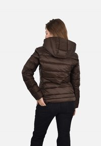 Hnedá prešívaná bunda s priliehavou kapucňou, prešívanou textúrou a kontrastnými vertikálnymi pruhmi. Neformálny dizajn, skombinovaná s tmavými džínsami.