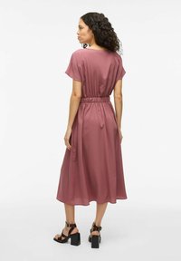 Mauve obleka srednje dolžine s kratkimi rokavi, elastičnim pasom in gladko teksturo. Črne masivne sandale na peto dopolnjujejo obleko.