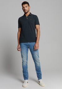 Donkerblauwe polo met een kraag, gecombineerd met lichtblauwe versleten jeans en witte sneakers. Heeft een klein logo op de borst.