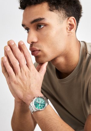 Giovane uomo con capelli corti e ricci che indossa un orologio da polso in argento con quadrante verde, poggiando il mento sulle mani con una camicia dai colori neutri.