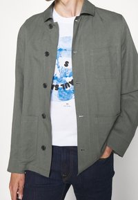 Šedá bavlněná bunda s límcem, zapínáním na knoflíky a n�áprsní kapsou. Nositá přes bílý grafikou potiskovaný tričko a tmavě modré džíny.