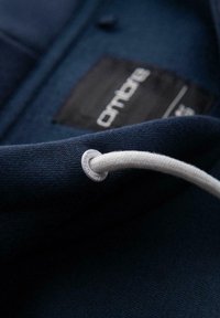 Granatowa tkanina z białym sznurkiem przewleczonym przez metalowe oczko; rozmyta czarna metka odzieżowa z nazwą marki w białym tekście.