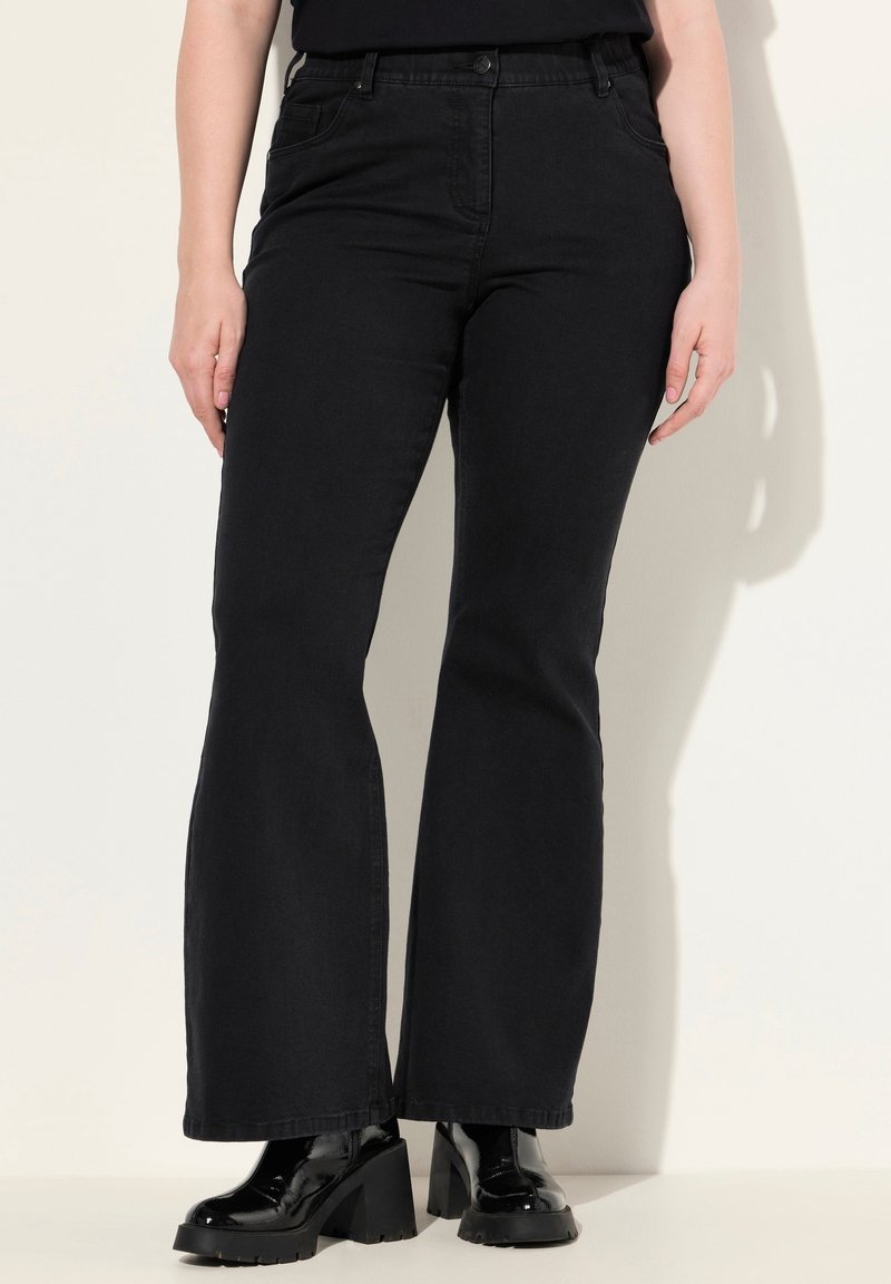 Schwarze Schlagjeans aus Denim, mit hoher Taille, Fronttaschen und klassischem Fünf-Taschen-Design. Kombiniert mit klobigen schwarzen Stiefeletten mit Absatz.