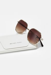 Brown gradient sunglasses with a square shape, gold metal frame, and decorative accents on the temples, resting on a white case embossed with "Michael Kors."