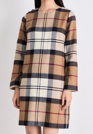 Kobieta w sukience z długim rękawem, sięgającej do kolan, w dużą kratę w kolorach beżu, czerni, bieli i czerwieni.