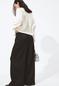 Donna con capelli scuri che indossa un maglione bianco e pantaloni neri larghi, mentre tiene una borsa bianca con macchie nere su uno sfondo bianco.