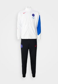Tracksuit med en hvid lynlåsjakke med blå ærmekanter, rødt Nike-logo og sorte bukser med blåt cirkulært logo og røde detaljer.