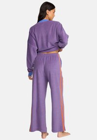 Conjunto de loungewear a rayas moradas que incluye una blusa corta y pantalones de pernera ancha. Destacado con rayas laterales naranjas y cinturilla elástica. Material suave.